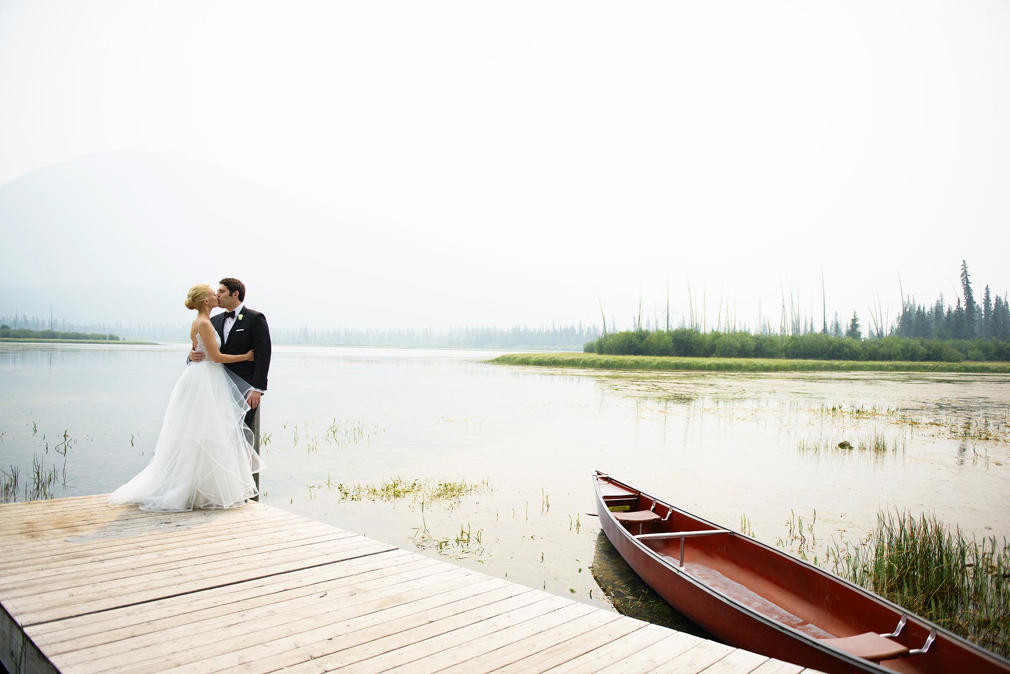 Couple on a wooden dock beside a red canoe on a misty lake near Banff