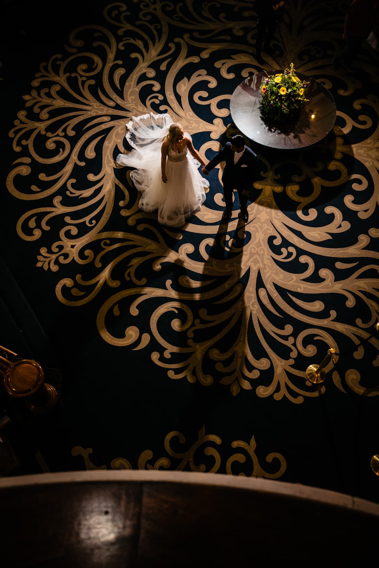 Bride and groom crossing a gold-patterned hotel ballroom floor, seen from the balcony above