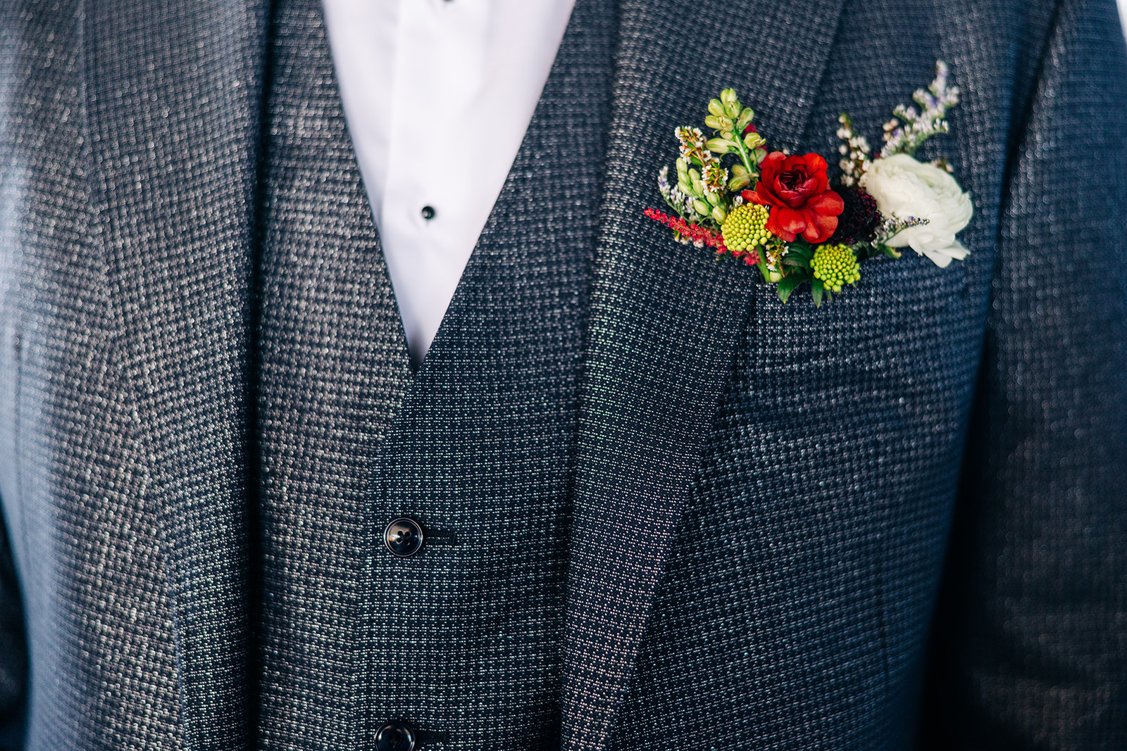 Groom's floral pocket boutonniere of red roses, ranunculus and greenery on a textured navy suit