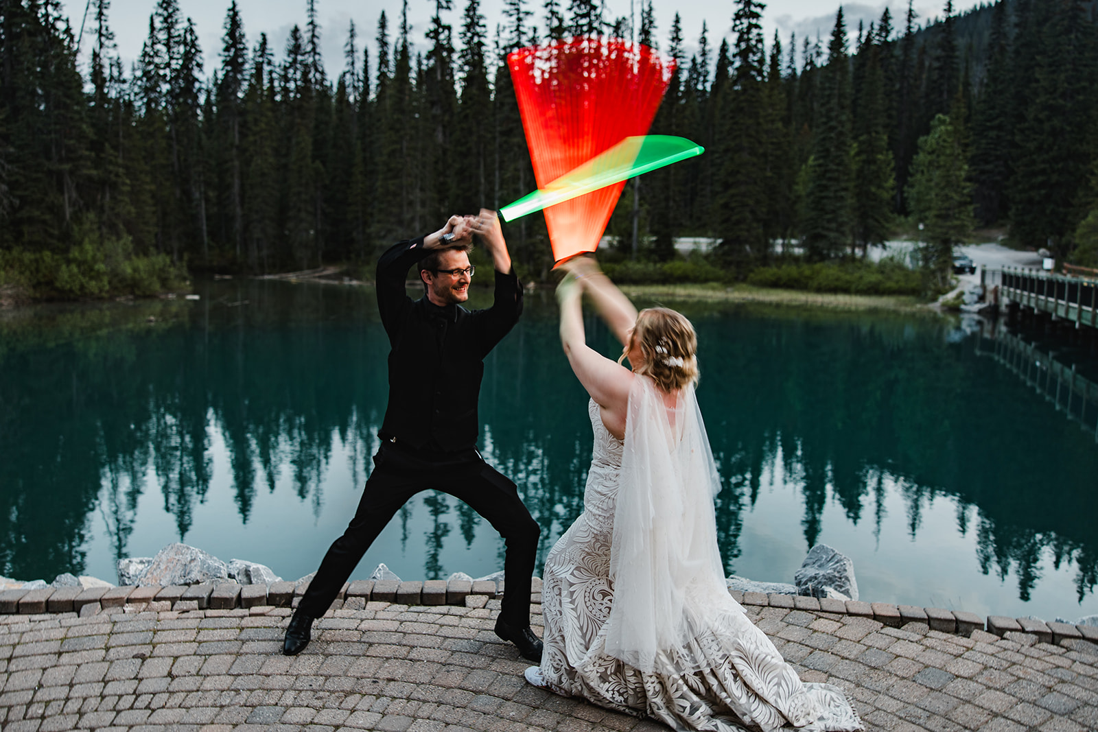 Bride and groom duelling with glowing sabres on the shore of Emerald Lake at dusk