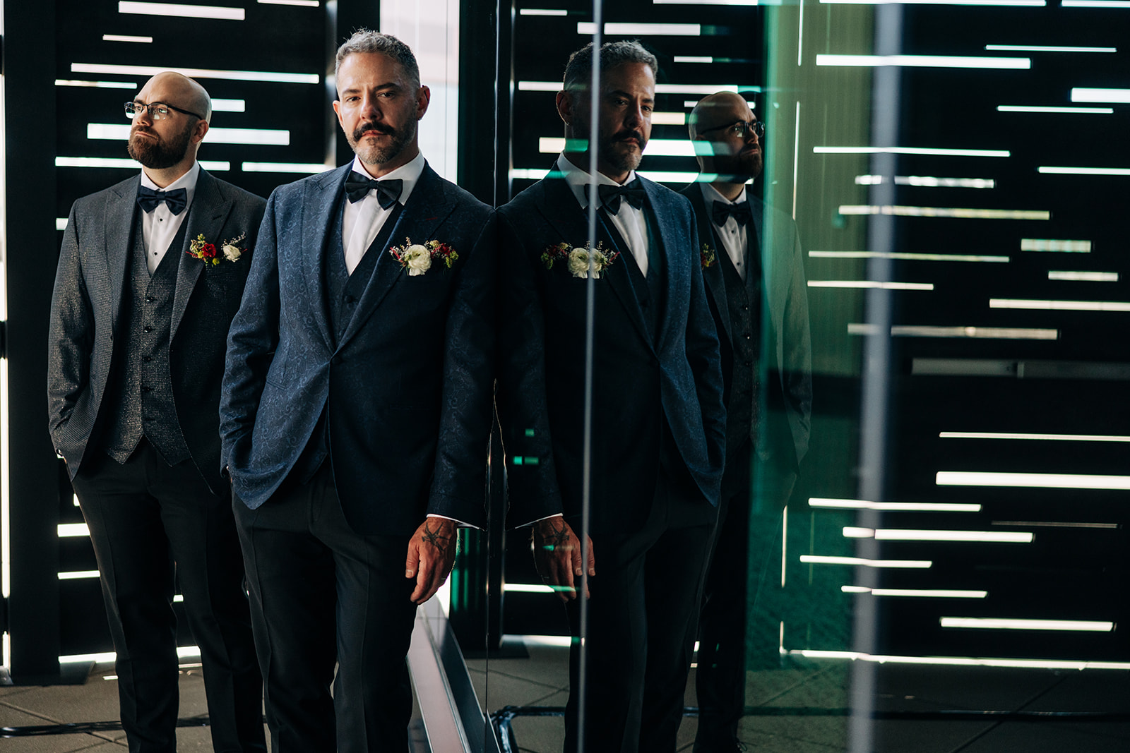 Two grooms reflected in glass beneath glowing light bars at their Stampede Park wedding in Calgary